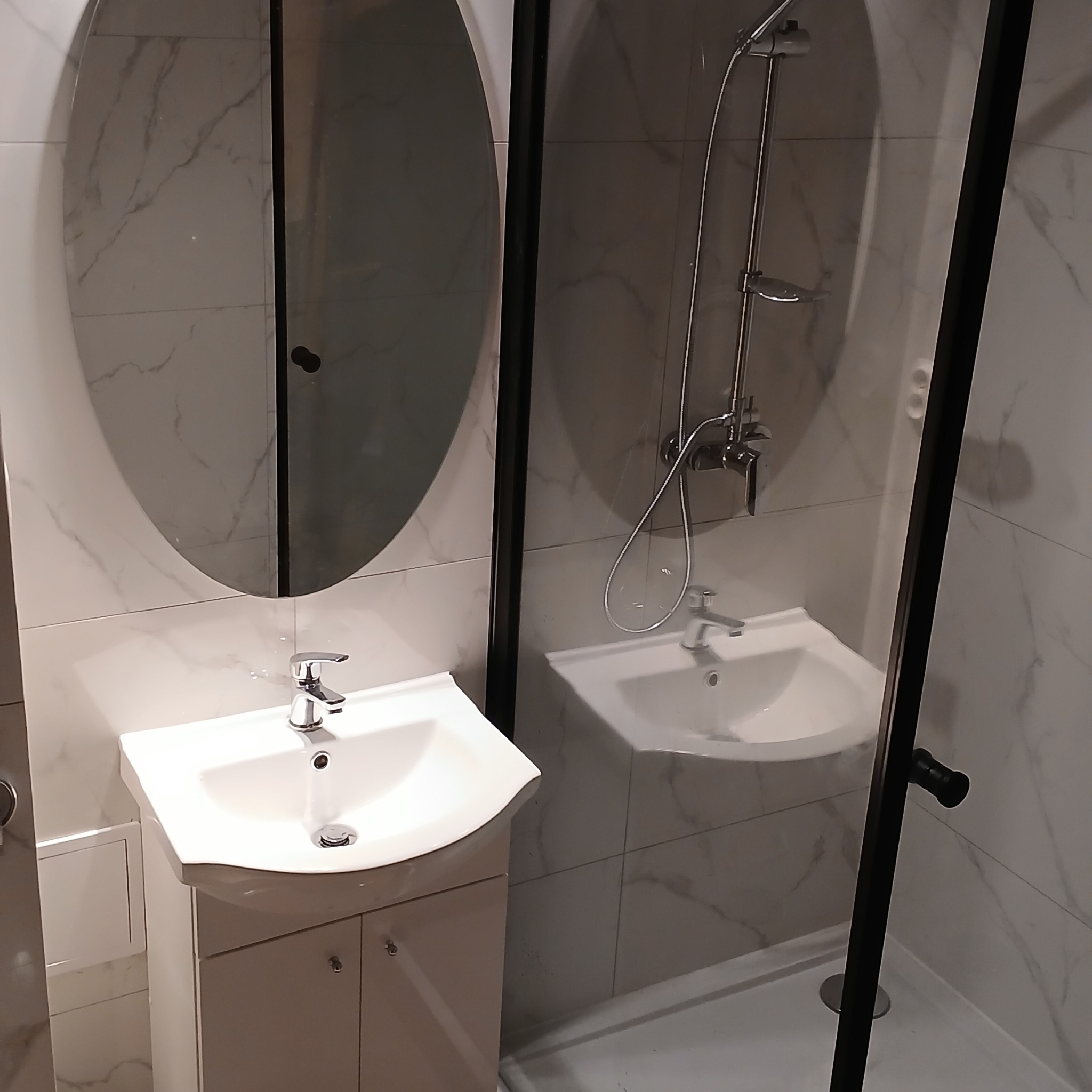 Nowoczesna łazienka z owalnym lustrem, białą umywalką na szafce i kabiną prysznicową z czarną ramą, wykończona płytkami imitującymi marmur.