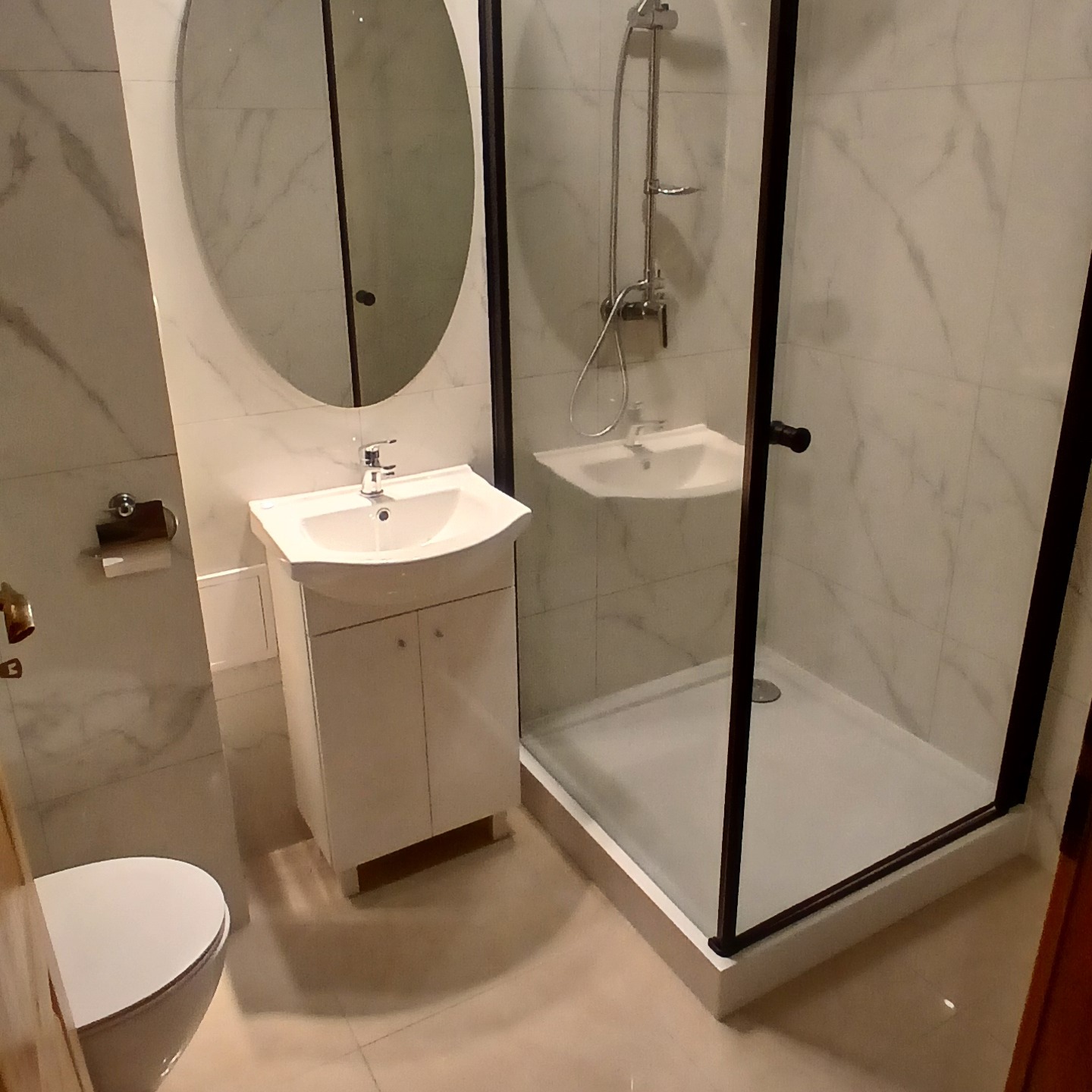 Nowoczesna łazienka z kabiną prysznicową z czarnymi ramami, owalnym lustrem nad białą szafką z umywalką i wiszącą toaletą, wykończona białymi płytkami imitującymi marmur.