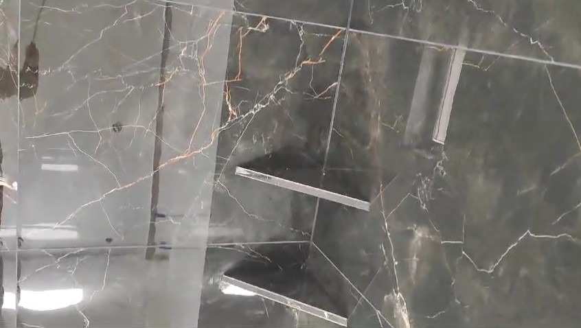 Ściana łazienki wyłożona ciemnymi, błyszczącymi płytkami imitującymi marmur z wyraźnym żyłowaniem w odcieniach bieli i miedzi. W rogu ściany zamontowane dwie półki z metalowymi wspornikami.