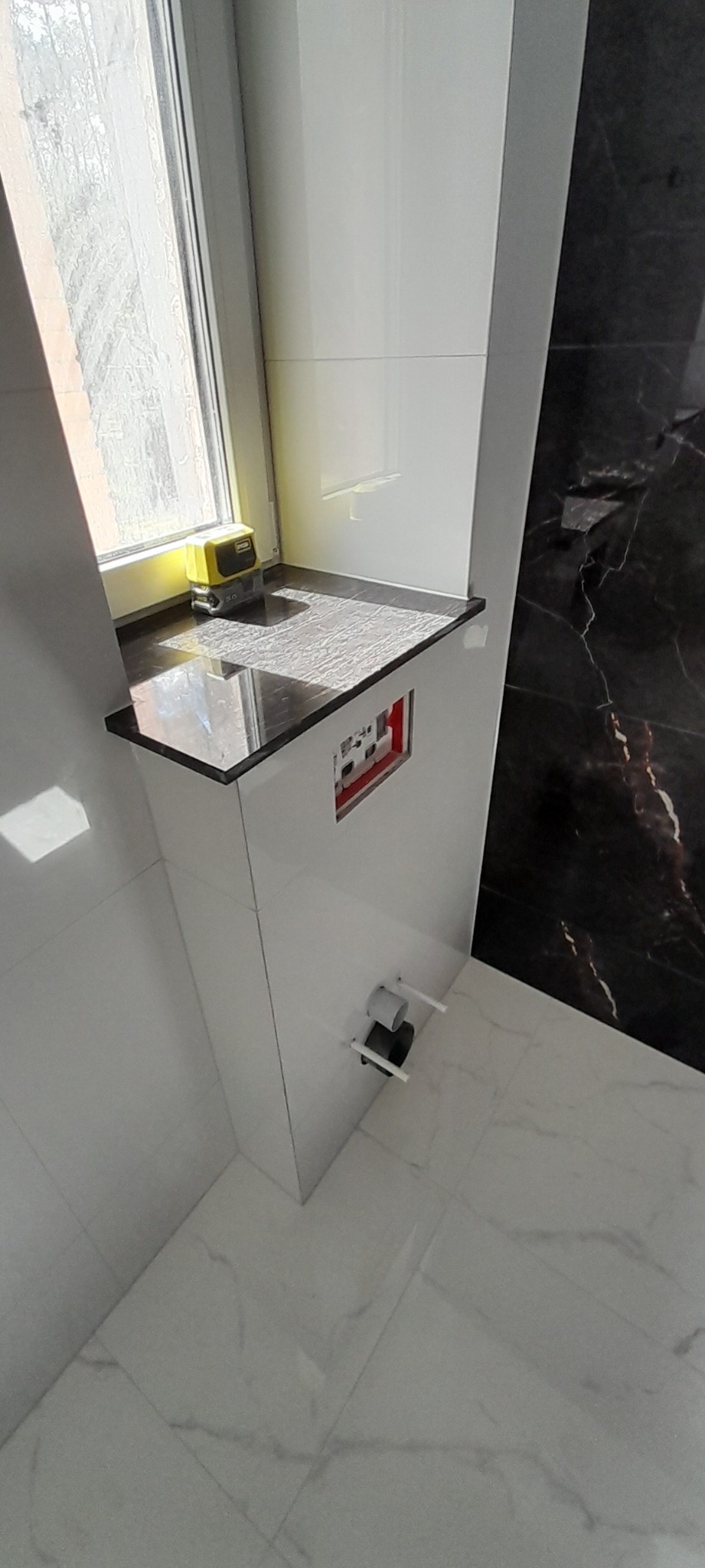 Biała, nowoczesna łazienka z czarnym blatem pod oknem, na którym leży żółty laser krzyżowy, widoczne przyłącza do armatury i marmurowe płytki.