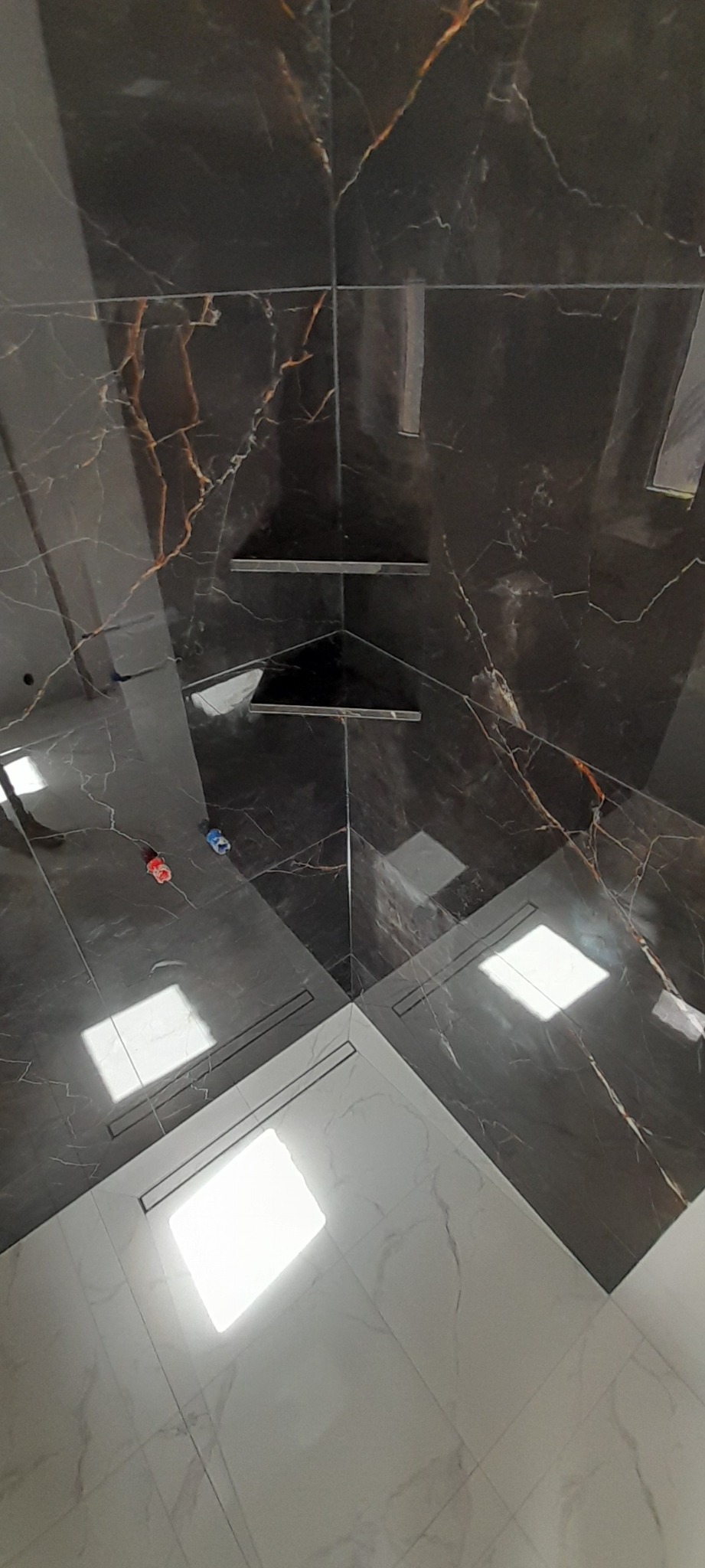 Ukończona łazienka z czarno-złotymi płytkami imitującymi marmur na ścianach i podłodze, w rogu wbudowane półki, widoczne odpływy liniowe w podłodze, perspektywa z góry.