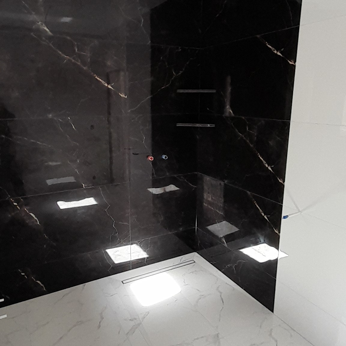 Częściowo wykończona kabina prysznicowa z czarnymi płytkami imitującymi marmur na ścianach i jasnymi płytkami na podłodze, widoczne przygotowane punkty wodne i odpływ liniowy.