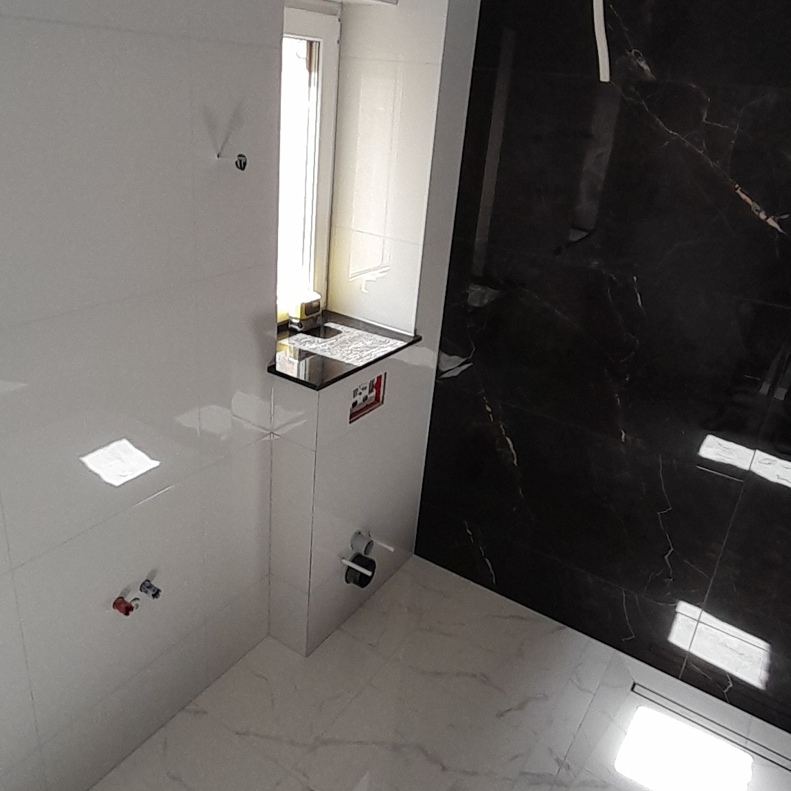 Nowoczesna łazienka z czarno-białymi płytkami imitującymi marmur, biała zabudowa geberit z czarnym blatem pod oknem, widoczne przyłącza wodne i kanalizacyjne.