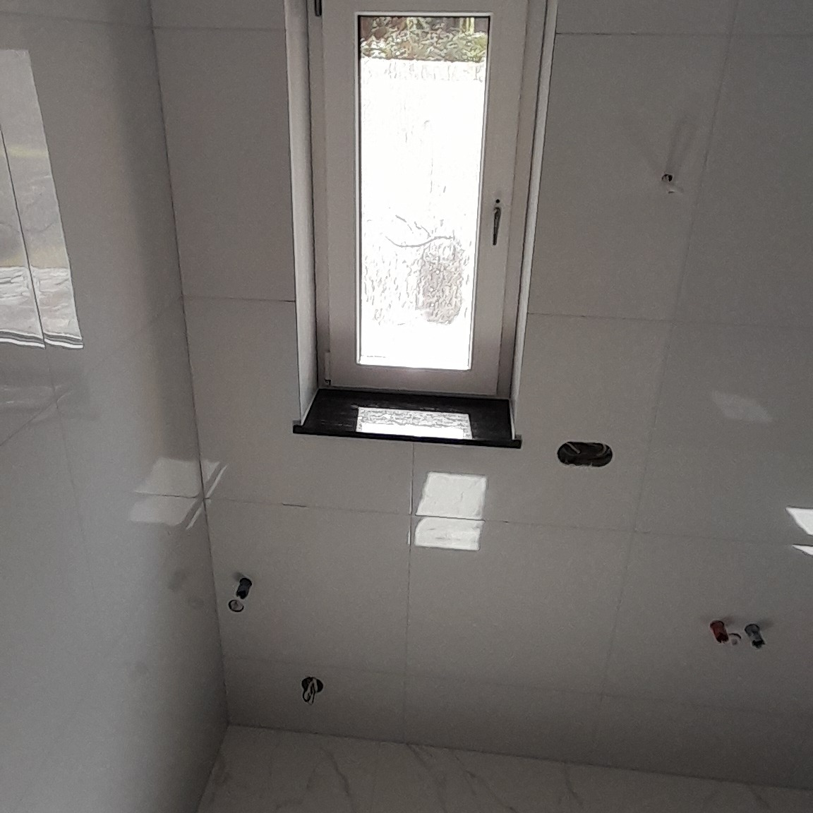 Białe płytki ścienne w łazience, widoczne otwory na instalacje wodno-kanalizacyjne i okno z czarnym parapetem.