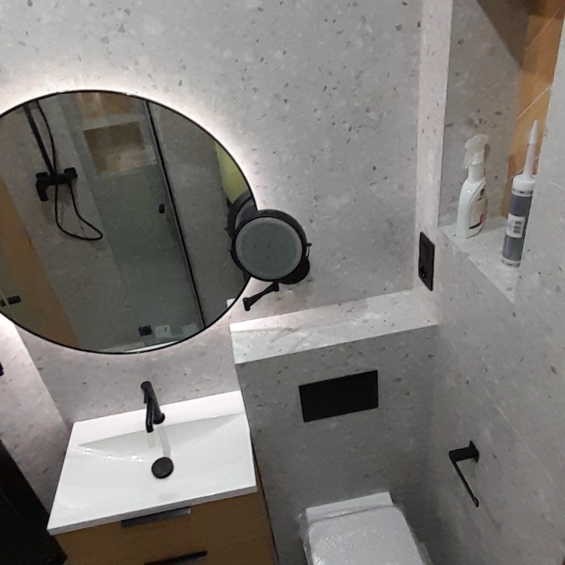 Nowoczesna łazienka z okrągłym lustrem podświetlanym LED, umywalką na szafce w kolorze drewna, toaletą i wnęką z kosmetykami, wykończona płytkami imitującymi lastryko.