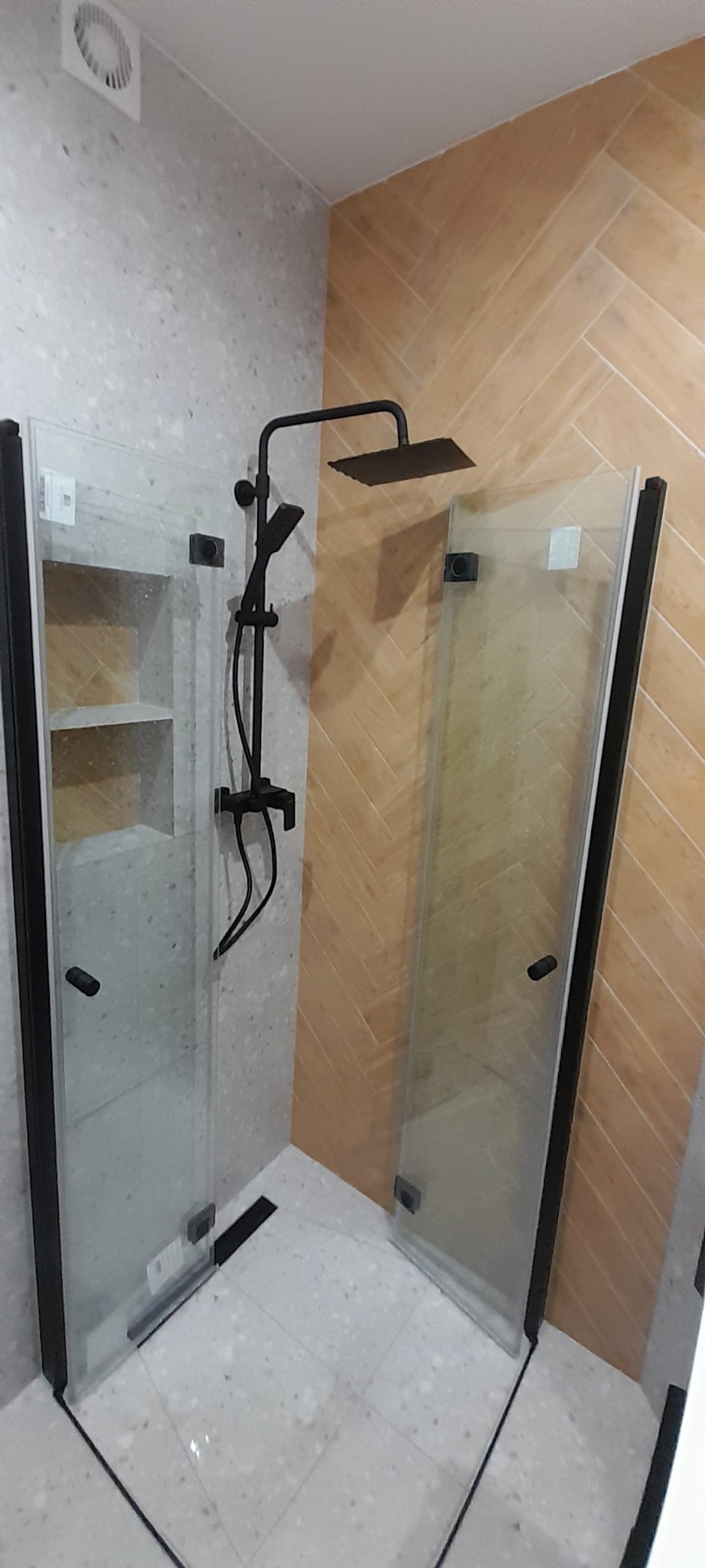 Nowoczesna kabina prysznicowa z czarnymi profilami i deszczownicą, ściany wyłożone płytkami imitującymi drewno i kamień, odpływ liniowy.