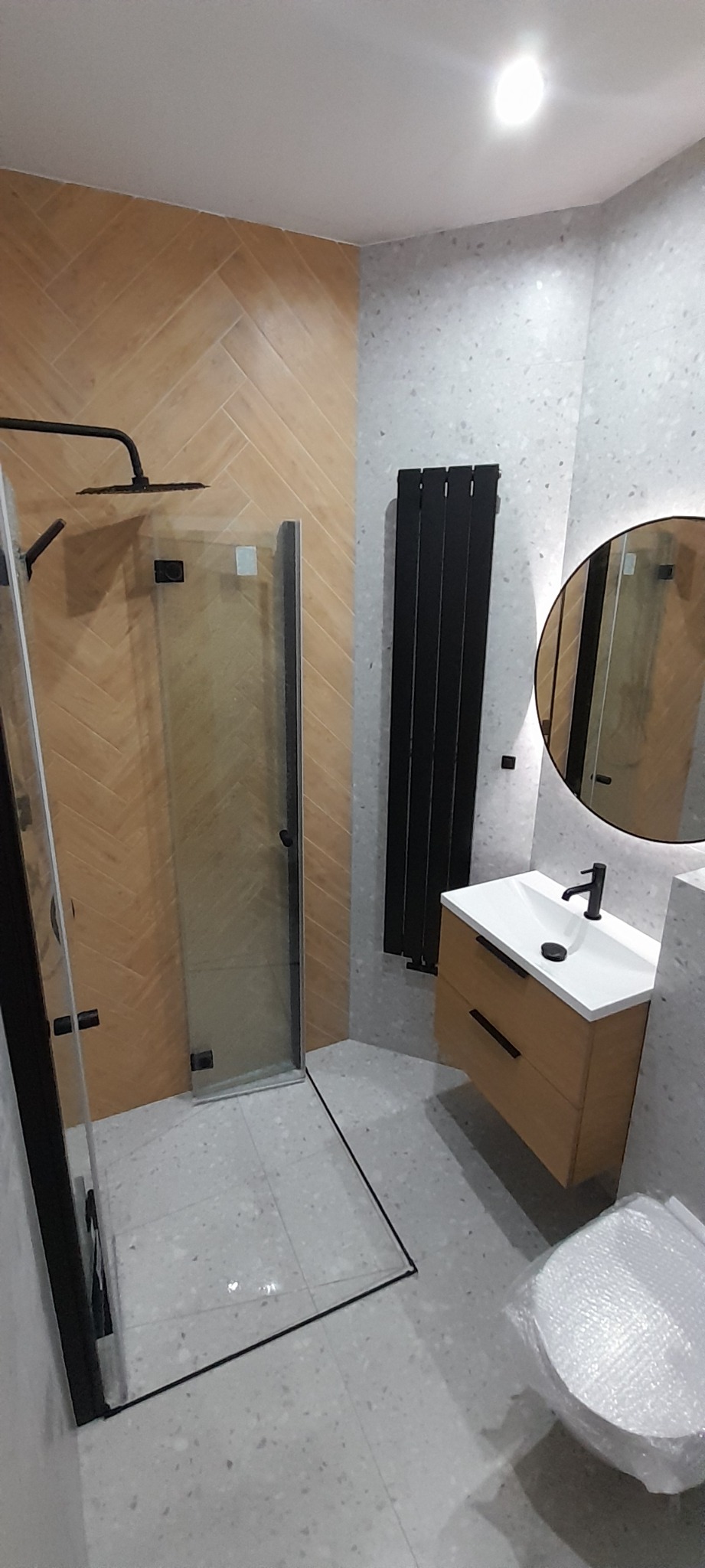 Nowoczesna łazienka z kabiną prysznicową typu walk-in z czarnymi profilami, drewnianą szafką pod umywalkę z czarną armaturą oraz grzejnikiem drabinkowym w kolorze czarnym, na ścianach płytki...