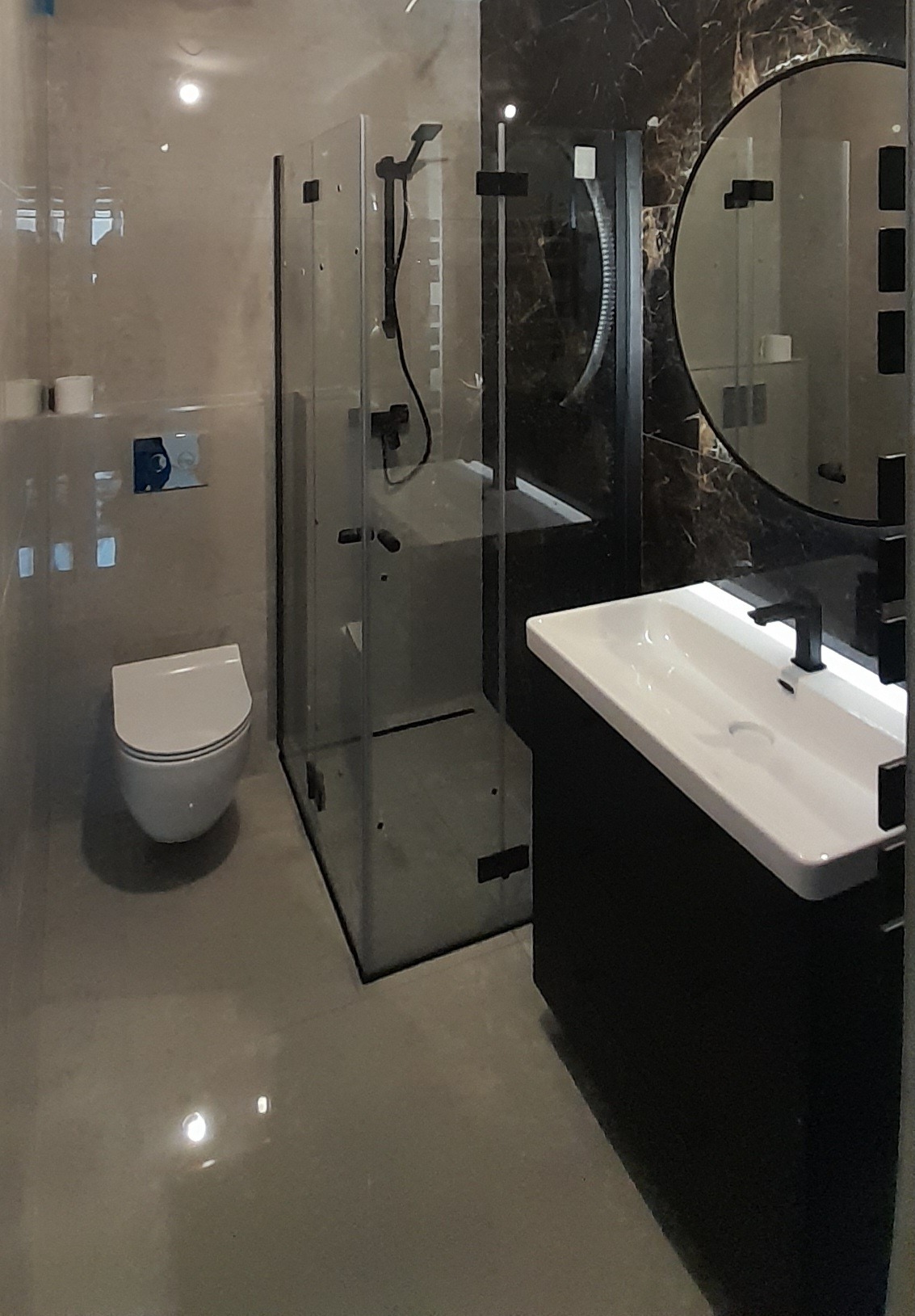 Nowoczesna łazienka z kabiną prysznicową typu walk-in, podwieszaną toaletą i umywalką nablatową na czarnej szafce, okrągłe lustro z czarną ramą na ścianie wykończonej marmurowym wzorem.