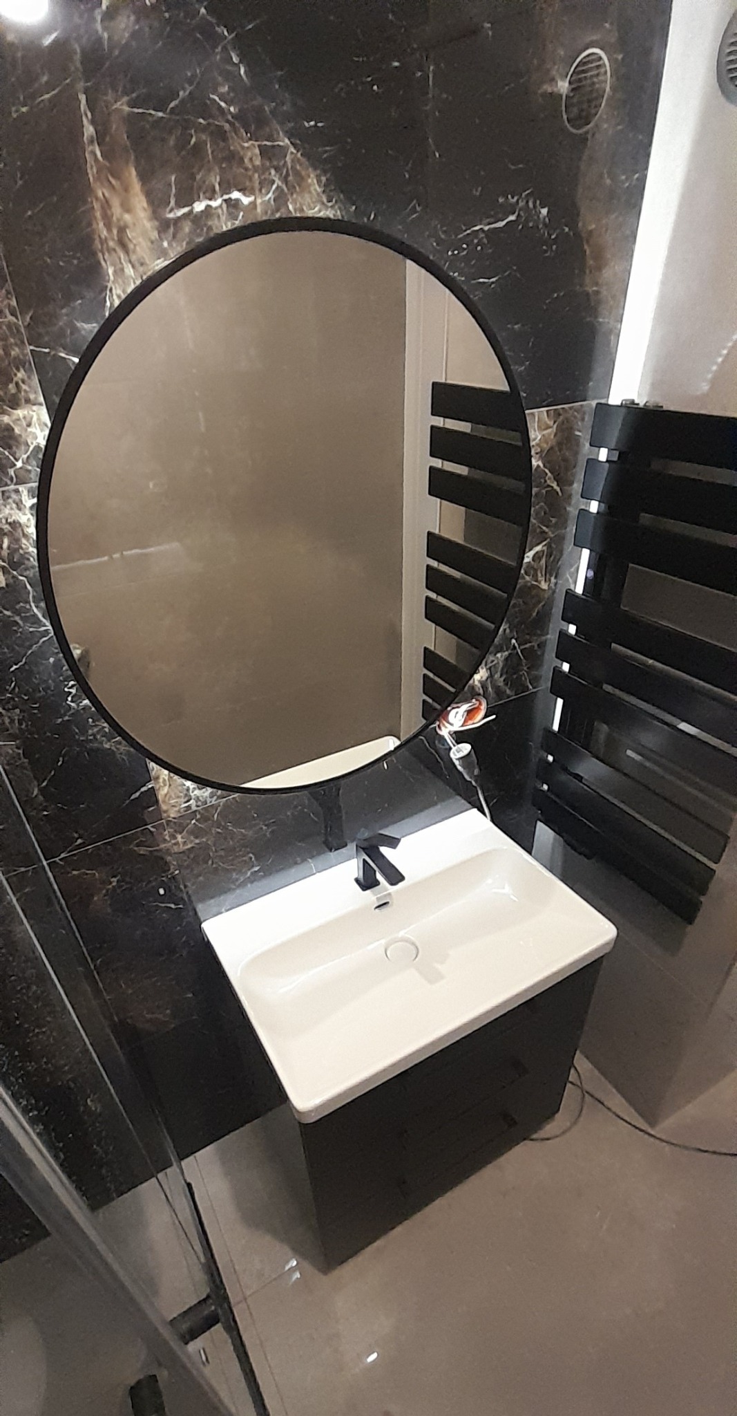 Nowoczesna łazienka z umywalką nablatową na czarnej szafce, okrągłe lustro z czarną ramą i grzejnik drabinkowy na tle ściany wyłożonej czarnym marmurem.