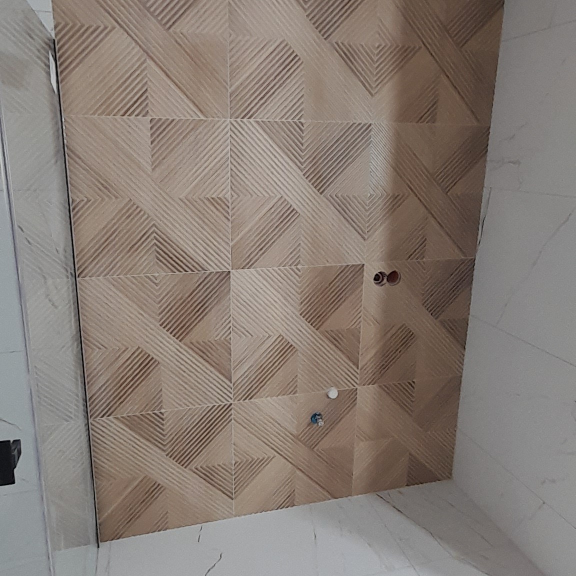 Ściana łazienki wyłożona beżowymi płytkami z geometrycznym wzorem, widoczne otwory na instalację wodną i fragment białej ściany z marmurkowym wzorem.
