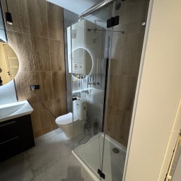 Usługi remontowe Maksym Kostiuk - Nowoczesna łazienka z kabiną prysznicową ze szklanymi drzwiami z czarnymi zawiasami, wiszącą toaletą, umywalką z szafką i okrągłym lustrem z podświetleniem LED, wykończona płytkami imitującymi drewno.