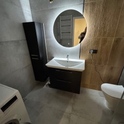 Usługi remontowe Maksym Kostiuk - Nowoczesna łazienka z szarymi płytkami, umywalką z szafką w kolorze czarnym, okrągłym lustrem z podświetleniem LED i wiszącą toaletą; w tle widoczny grzejnik drabinkowy i pralka z otwartymi...