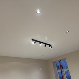 Usługi remontowe Maksym Kostiuk - Biały sufit z zamontowanym czarnym reflektorem szynowym z sześcioma punktami świetlnymi i kilkoma pojedynczymi, okrągłymi lampami wpuszczanymi. Widoczne dwie ściany pomalowane na jasny, neutralny...