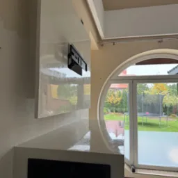 Ściana z dekoracyjną, błyszczącą gładzią w odcieniach bieli i beżu, z zamontowanym uchwytem na telewizor, widok na ogród przez duże, okrągłe okno.