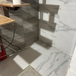 Usługi remontowe Maksym Kostiuk - Fragment łazienki w trakcie remontu: ułożone płytki imitujące marmur na ścianach i podłodze, widoczne przewody i prowizoryczne półki, rolka papieru na górnej półce.