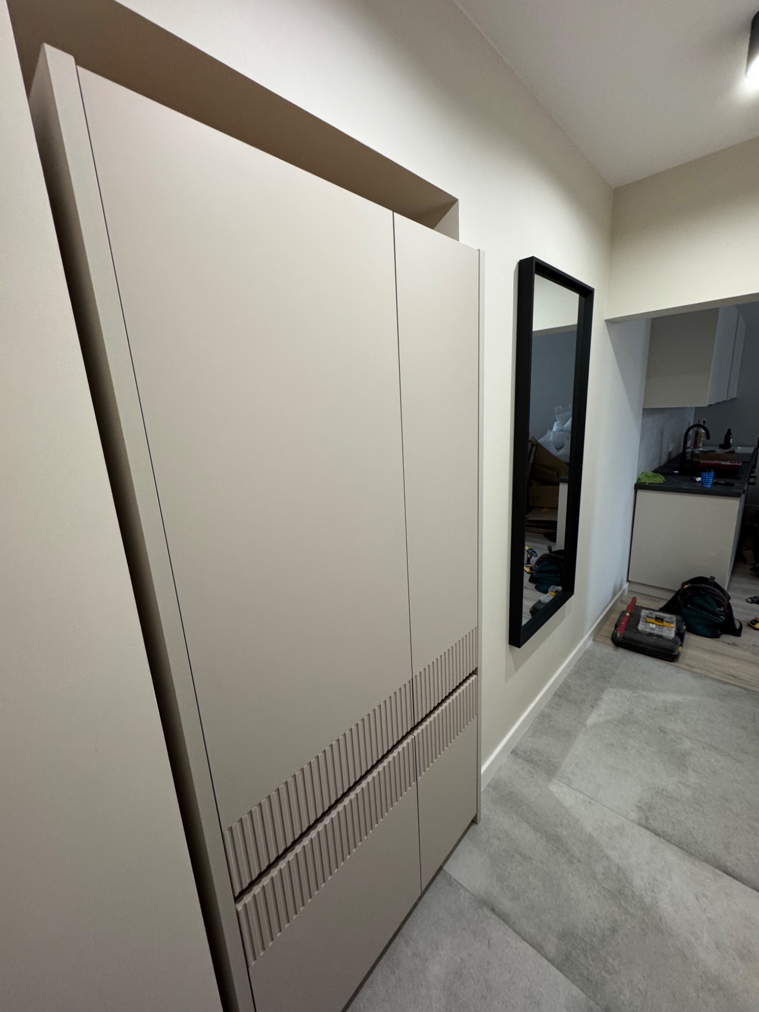 Beżowa szafa wnękowa z dekoracyjnymi frezami na frontach, czarne lustro w wąskiej ramie na ścianie, szare płytki podłogowe, widok na fragment kuchni w tle.