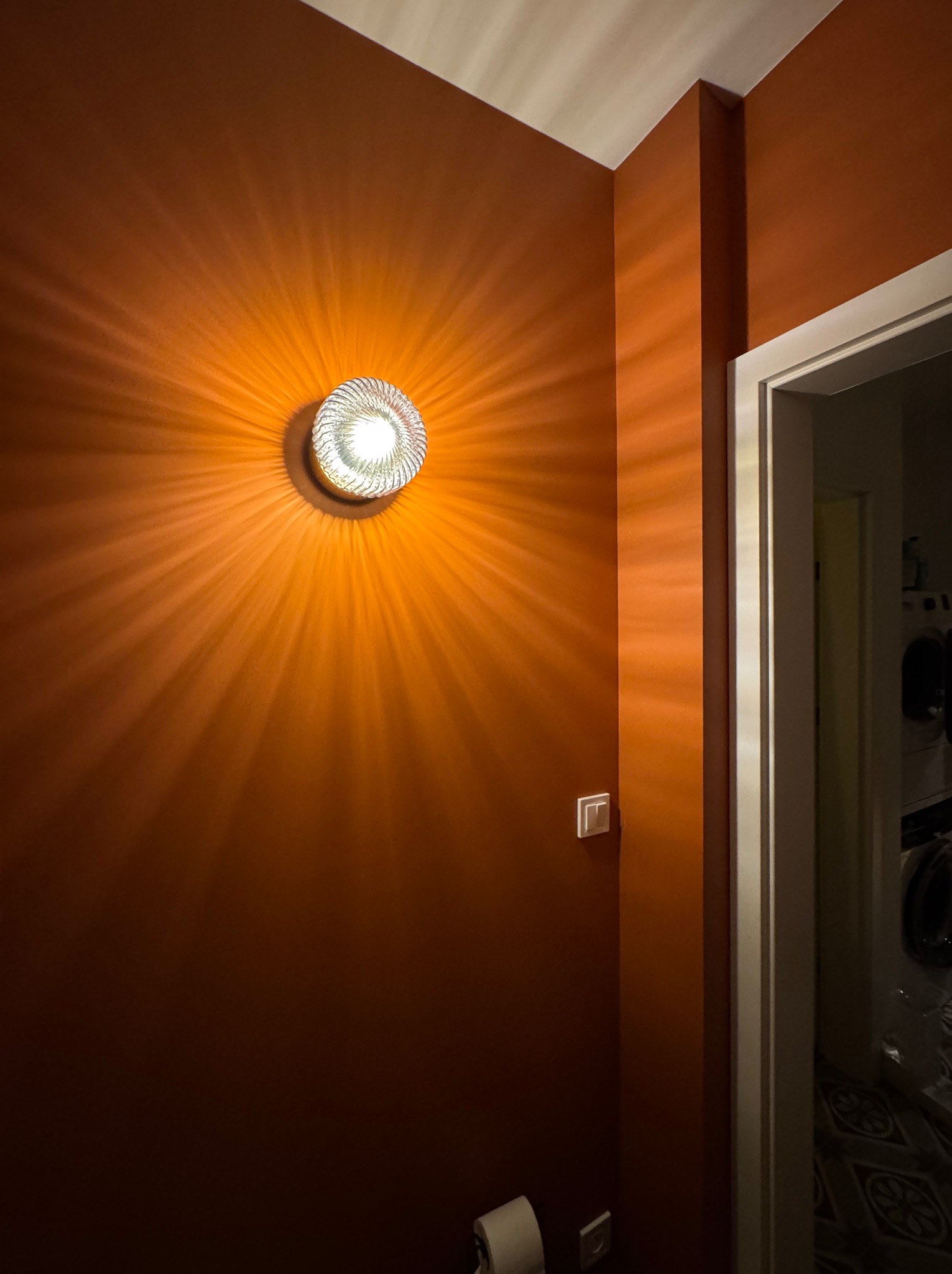 Ściana w odcieniach terakoty oświetlona dekoracyjnym kinkietem z efektem promienistego światła, widoczny fragment ramy drzwiowej i włącznik elektryczny.