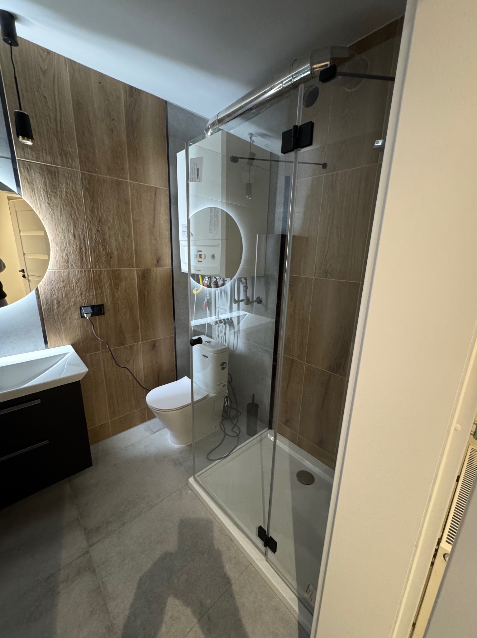 Nowoczesna łazienka z kabiną prysznicową ze szklanymi drzwiami z czarnymi zawiasami, wiszącą toaletą, umywalką z szafką i okrągłym lustrem z podświetleniem LED, wykończona płytkami imitującymi drewno.