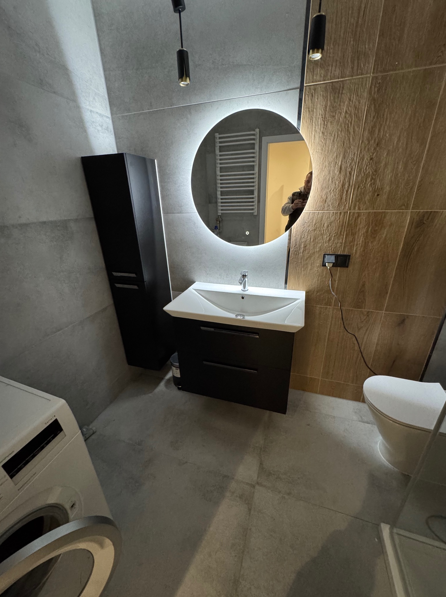 Nowoczesna łazienka z szarymi płytkami, umywalką z szafką w kolorze czarnym, okrągłym lustrem z podświetleniem LED i wiszącą toaletą; w tle widoczny grzejnik drabinkowy i pralka z otwartymi...