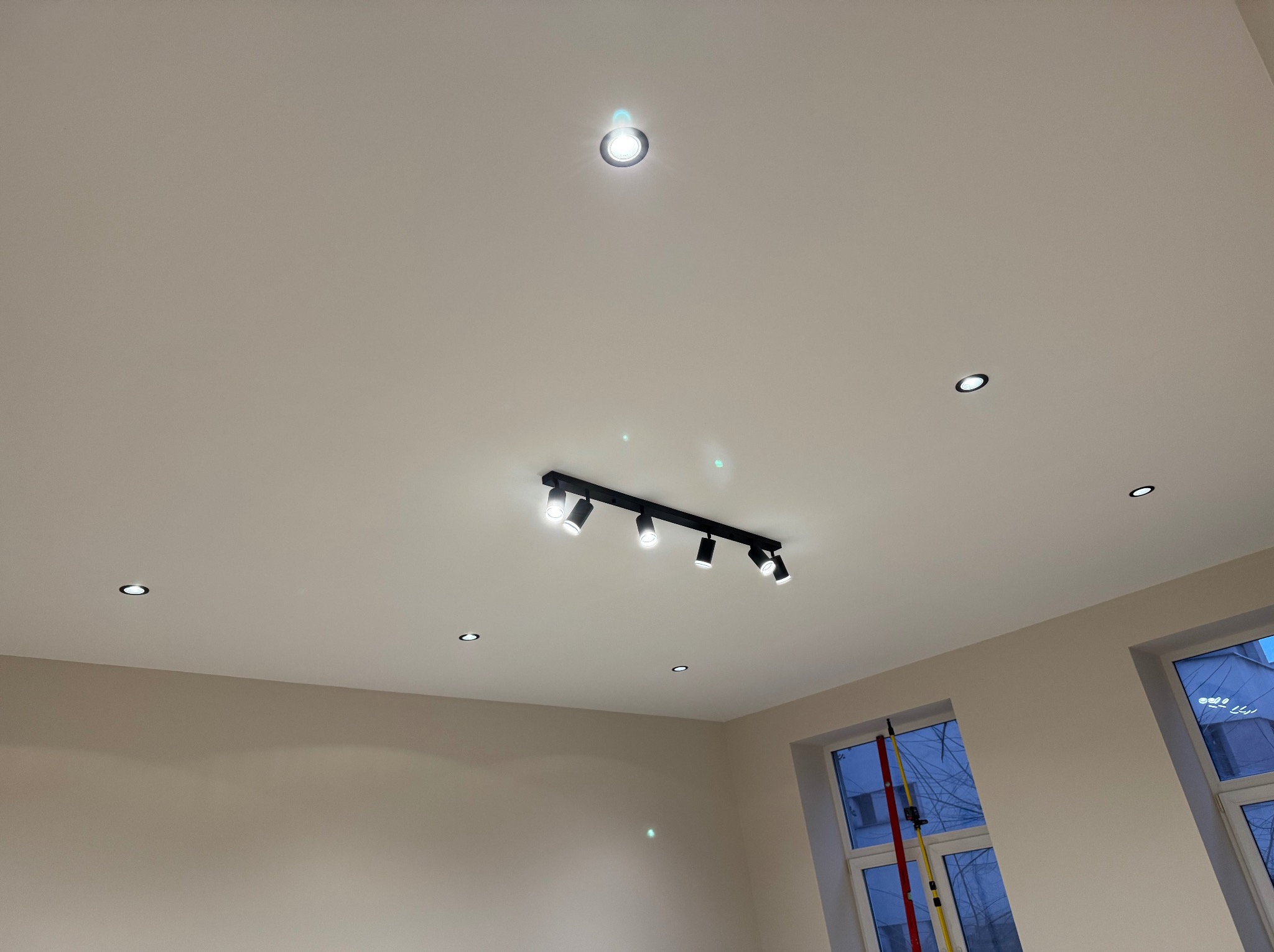 Biały sufit z zamontowanym czarnym reflektorem szynowym z sześcioma punktami świetlnymi i kilkoma pojedynczymi, okrągłymi lampami wpuszczanymi. Widoczne dwie ściany pomalowane na jasny, neutralny...