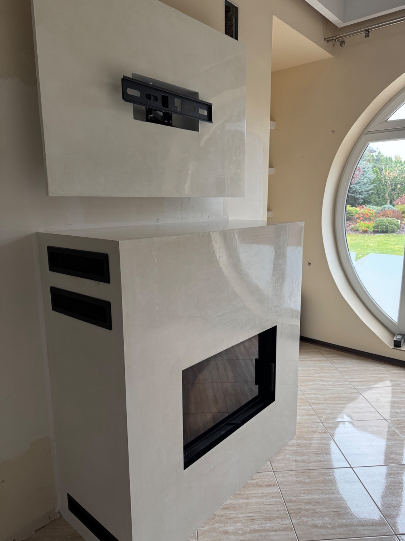 Nowoczesny kominek obłożony jasną, błyszczącą gładzią w salonie z okrągłym oknem i widokiem na ogród, z zamontowanym uchwytem na telewizor powyżej.