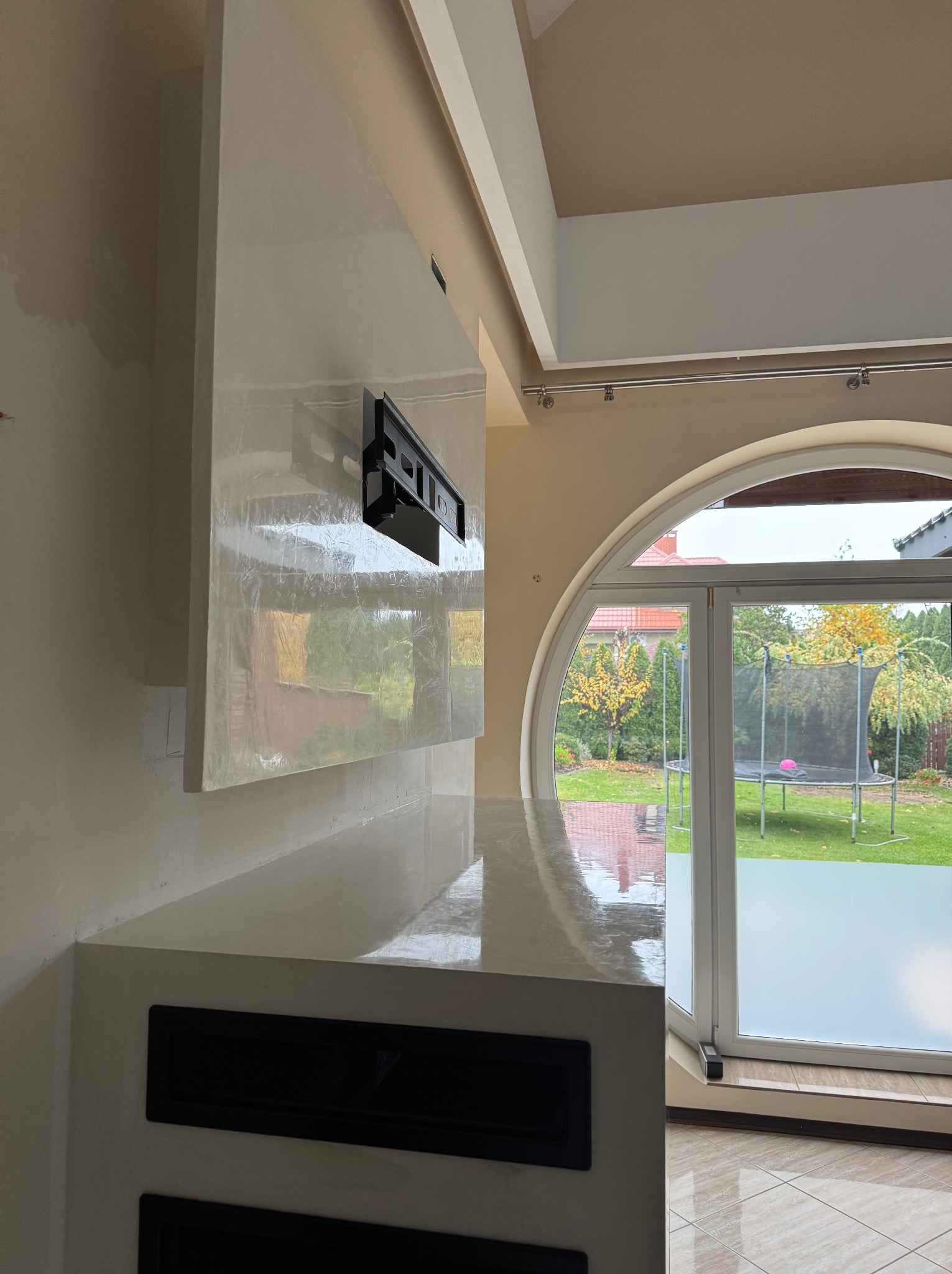 Ściana z dekoracyjną, błyszczącą gładzią w odcieniach bieli i beżu, z zamontowanym uchwytem na telewizor, widok na ogród przez duże, okrągłe okno.