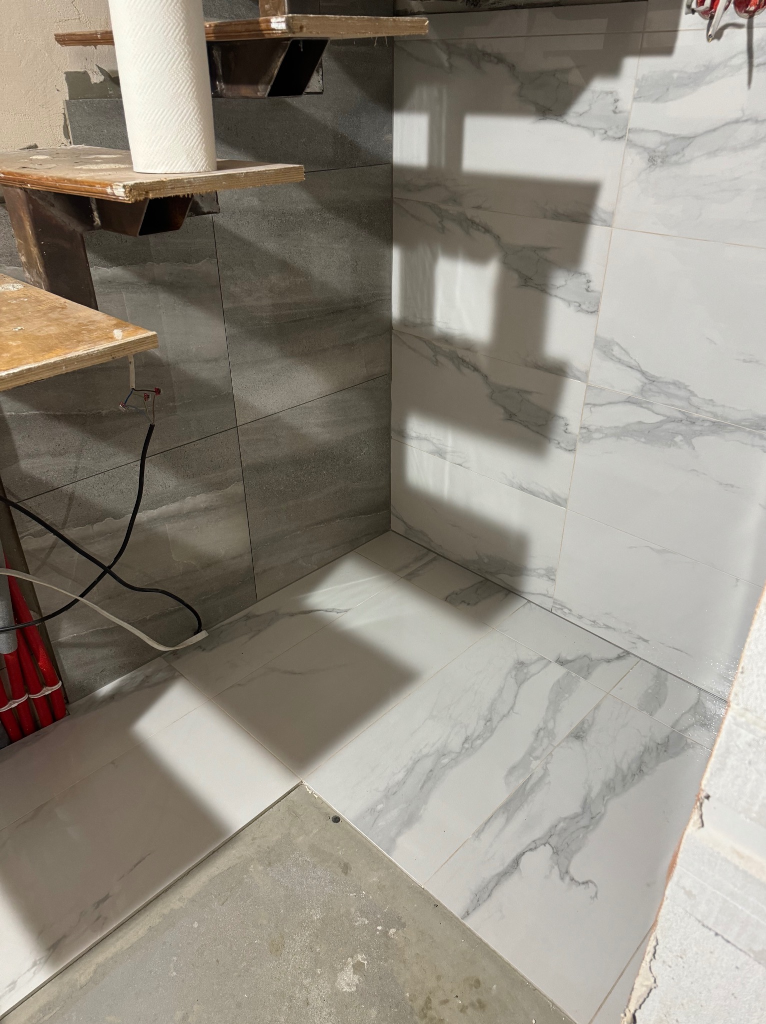 Fragment łazienki w trakcie remontu: ułożone płytki imitujące marmur na ścianach i podłodze, widoczne przewody i prowizoryczne półki, rolka papieru na górnej półce.