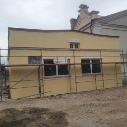 P.H.U KINA - Żółta elewacja budynku w trakcie prac wykończeniowych, widoczne rusztowania i przygotowane płyty izolacyjne przy oknach, w tle inny budynek.