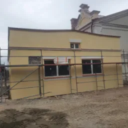 Żółta elewacja budynku w trakcie prac wykończeniowych, widoczne rusztowania i przygotowane płyty izolacyjne przy oknach, w tle inny budynek.