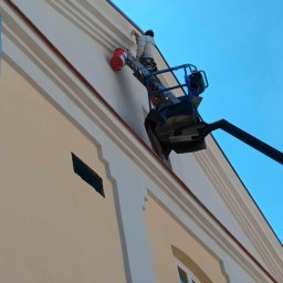 P.H.U KINA - Pracownik na podnośniku maluje elewację budynku z dekoracyjnymi elementami architektonicznymi, widoczny czerwony pojemnik z farbą.