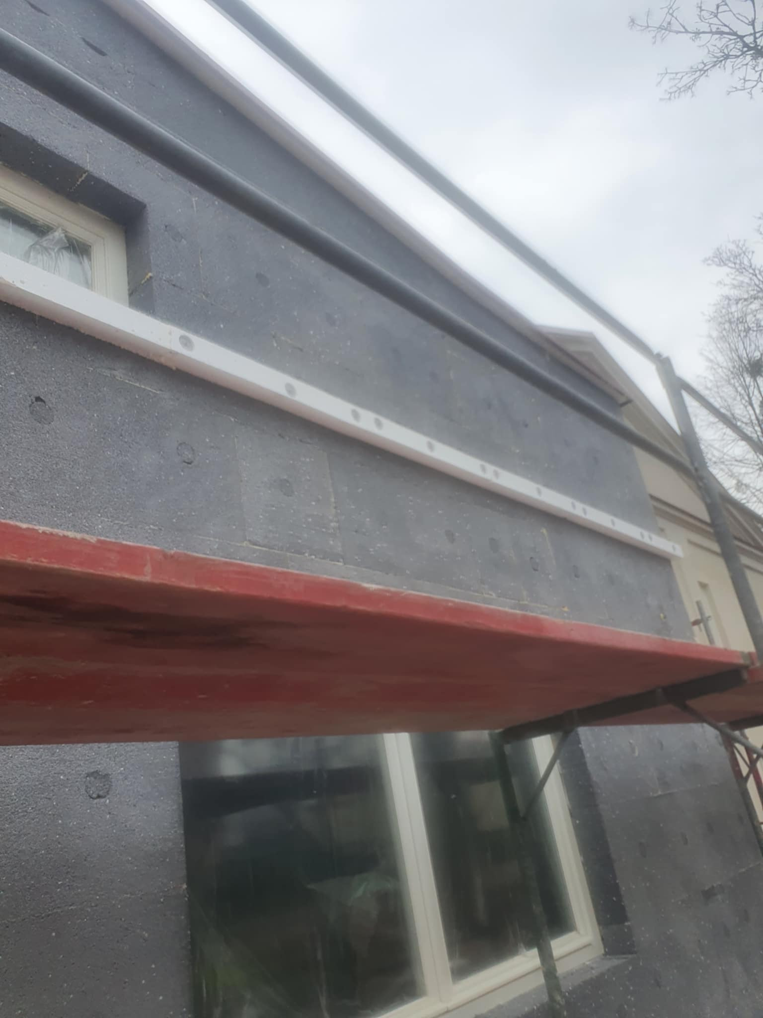 Szara elewacja budynku w trakcie ocieplania, widoczne płyty izolacyjne i rusztowanie z czerwonymi elementami, białe listwy dekoracyjne wokół okna.