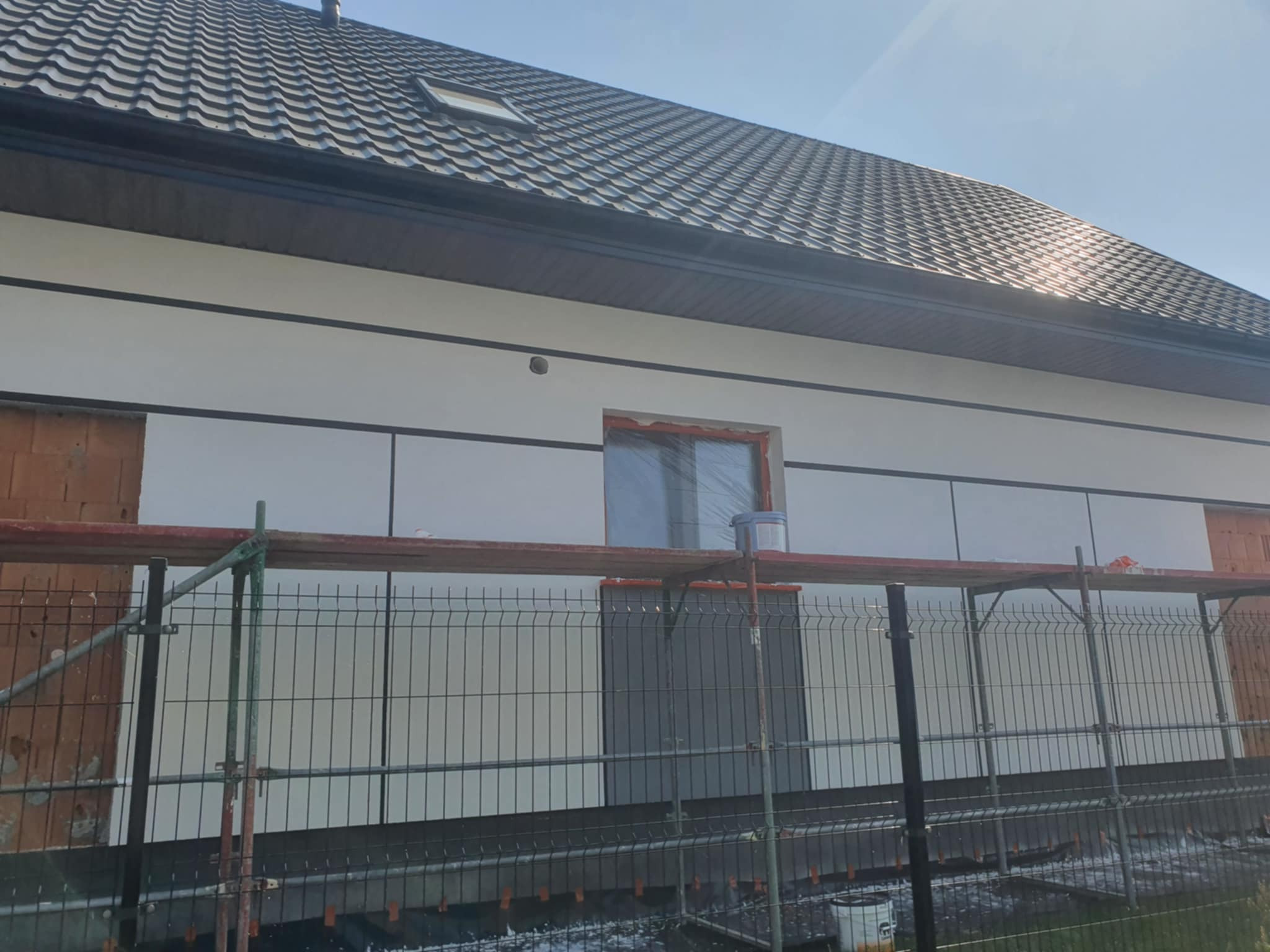 Fragment elewacji domu w trakcie prac wykończeniowych; widoczne rusztowania, częściowo otynkowana ściana i okno zabezpieczone folią.