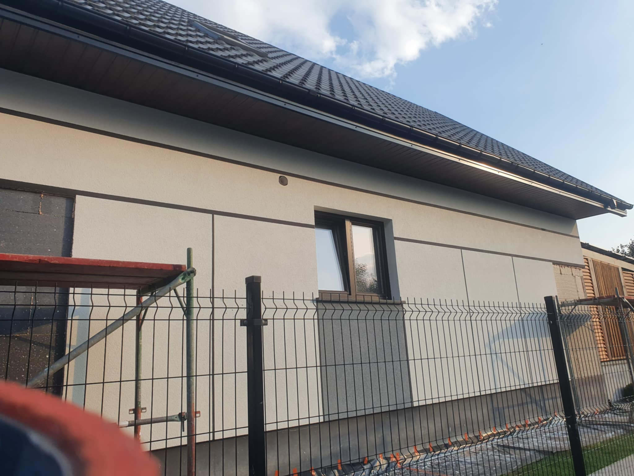 Fragment elewacji domu jednorodzinnego z widocznym oknem, wykończona jasnym tynkiem strukturalnym z ciemnymi akcentami, częściowo zasłonięta przez metalowe ogrodzenie panelowe i fragment rusztowania.