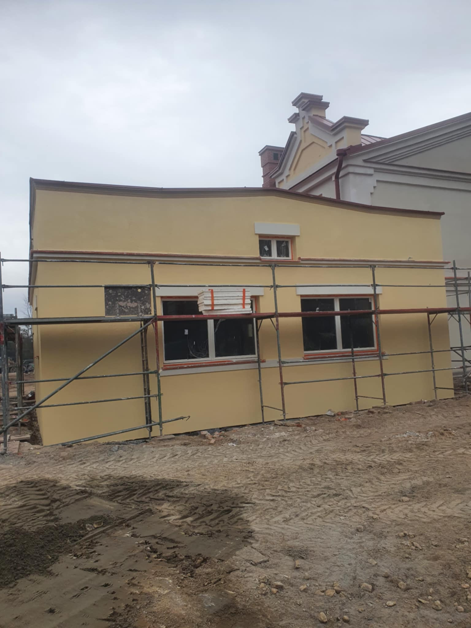 Żółta elewacja budynku w trakcie prac wykończeniowych, widoczne rusztowania i przygotowane płyty izolacyjne przy oknach, w tle inny budynek.
