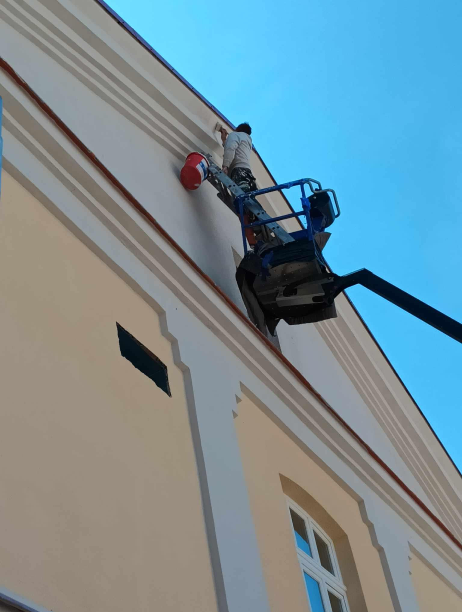 Pracownik na podnośniku maluje elewację budynku z dekoracyjnymi elementami architektonicznymi, widoczny czerwony pojemnik z farbą.