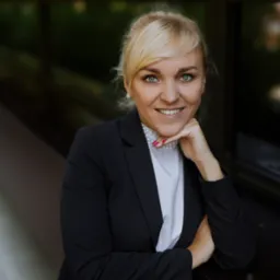 Uśmiechnięta blondynka w czarnym żakiecie i perłowym naszyjniku, oparta podbródkiem o dłoń, patrzy w obiektyw.