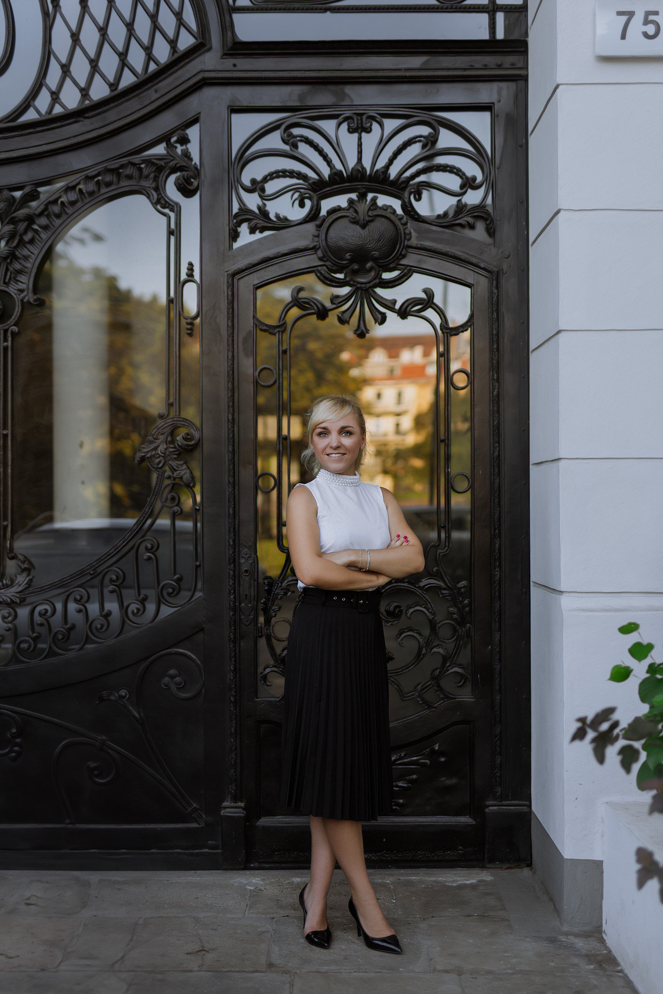 Uśmiechnięta blondynka w eleganckim stroju, stojąca na tle ozdobnych, czarnych drzwi z numerem 75 na budynku.