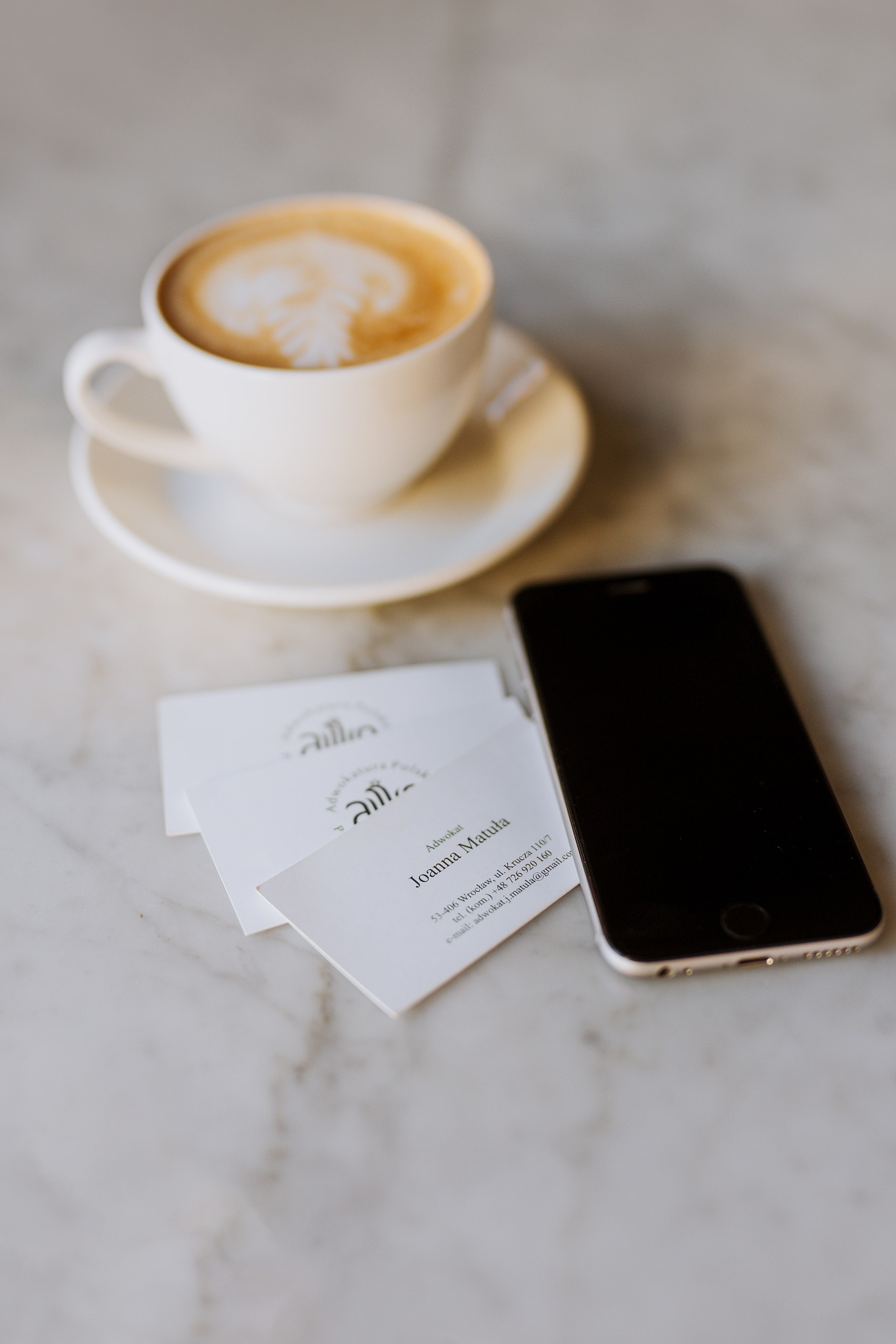Kawa latte w białej filiżance na marmurowym stole obok wizytówek adwokata Joanna Matuła i smartfona