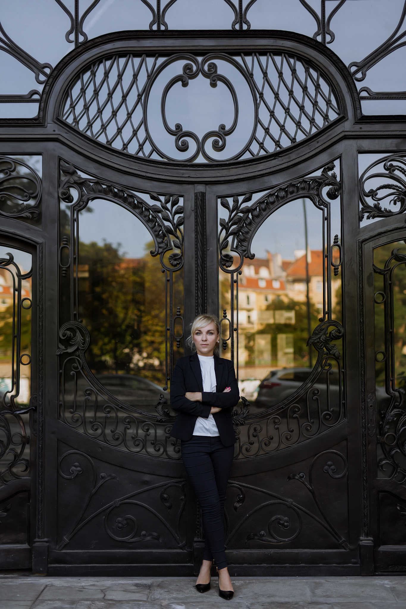 Kobieta w garniturze oparta o ozdobną, kutą bramę z widocznymi budynkami w tle.