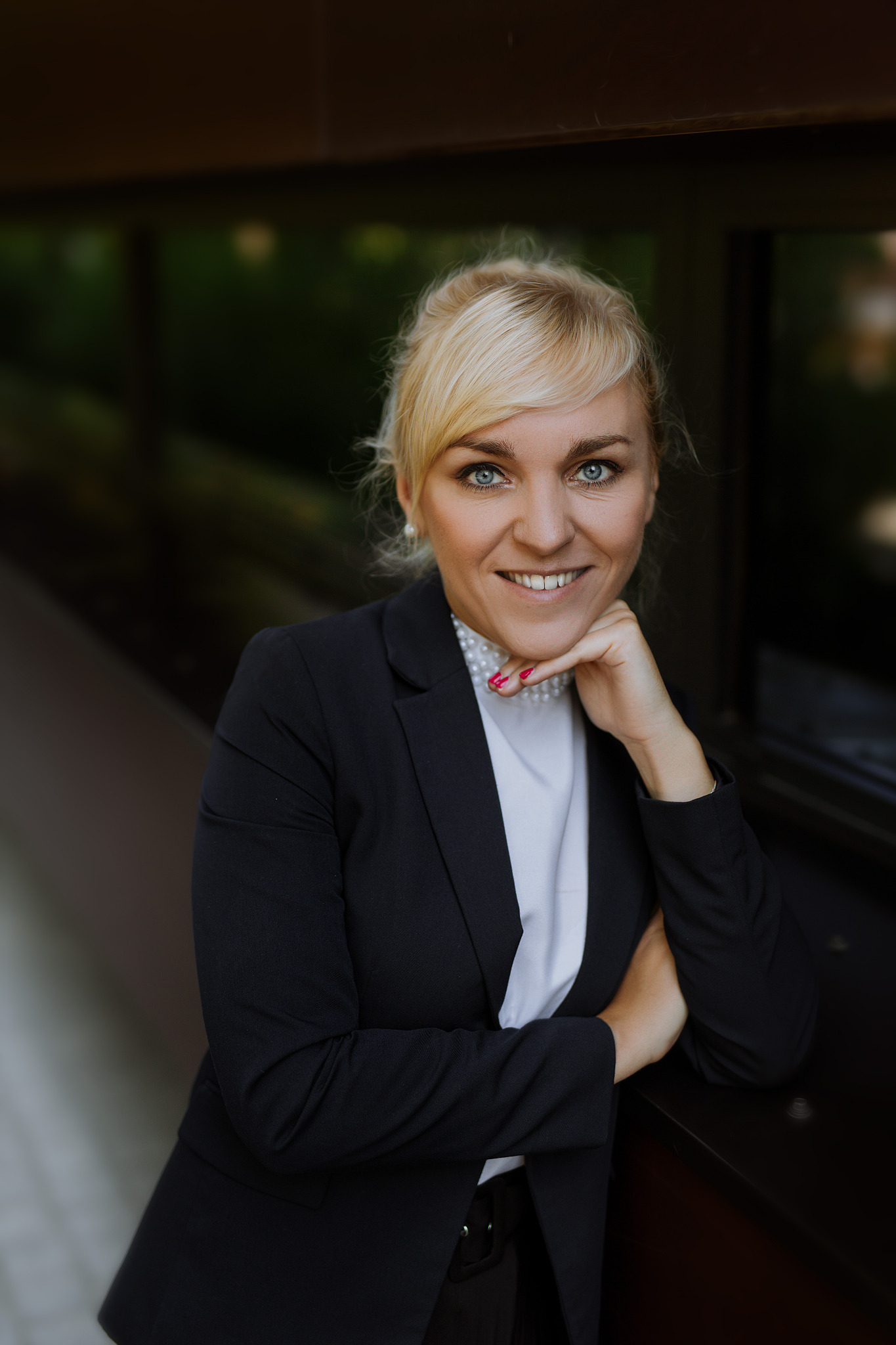 Uśmiechnięta blondynka w czarnym żakiecie i perłowym naszyjniku, oparta podbródkiem o dłoń, patrzy w obiektyw.