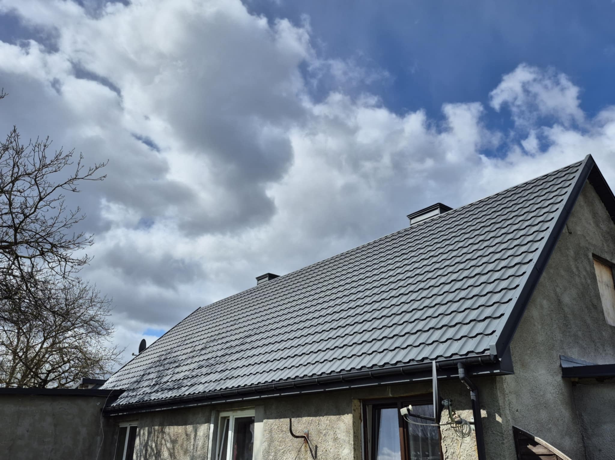 Dom z nowym, szarym dachem z blachodachówki modułowej, widoczne rynny i komin na tle pochmurnego nieba.