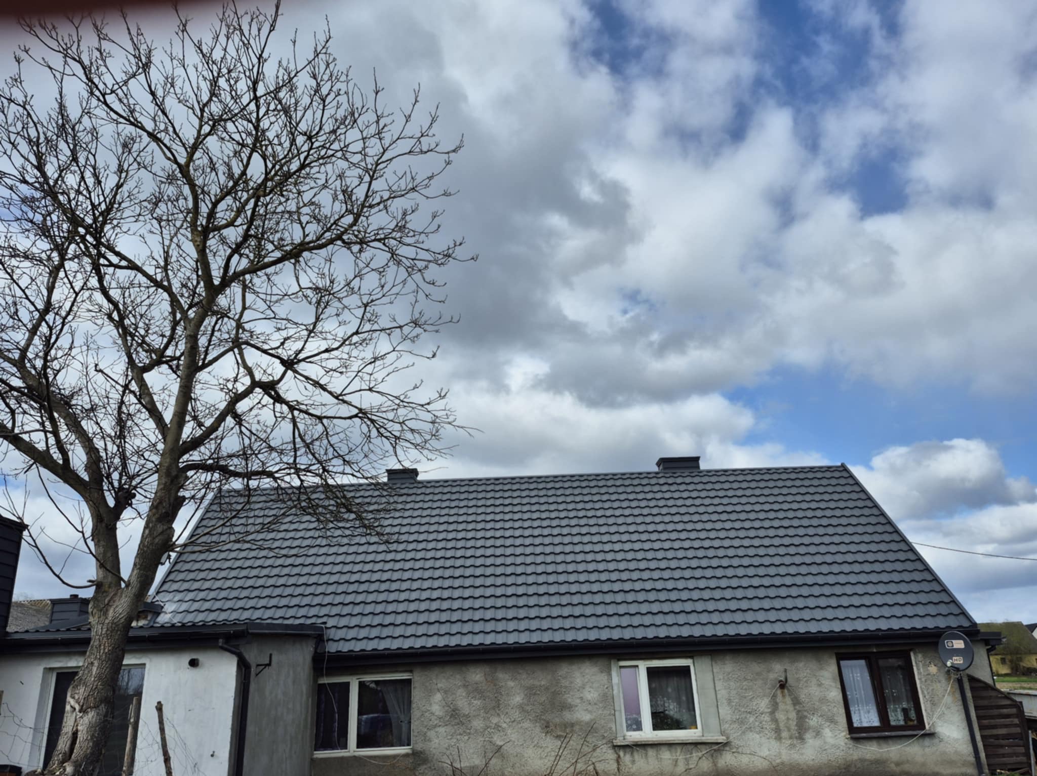Dom z nowym, grafitowym dachem z blachodachówki, widoczny komin i antena satelitarna, na pierwszym planie drzewo bez liści.