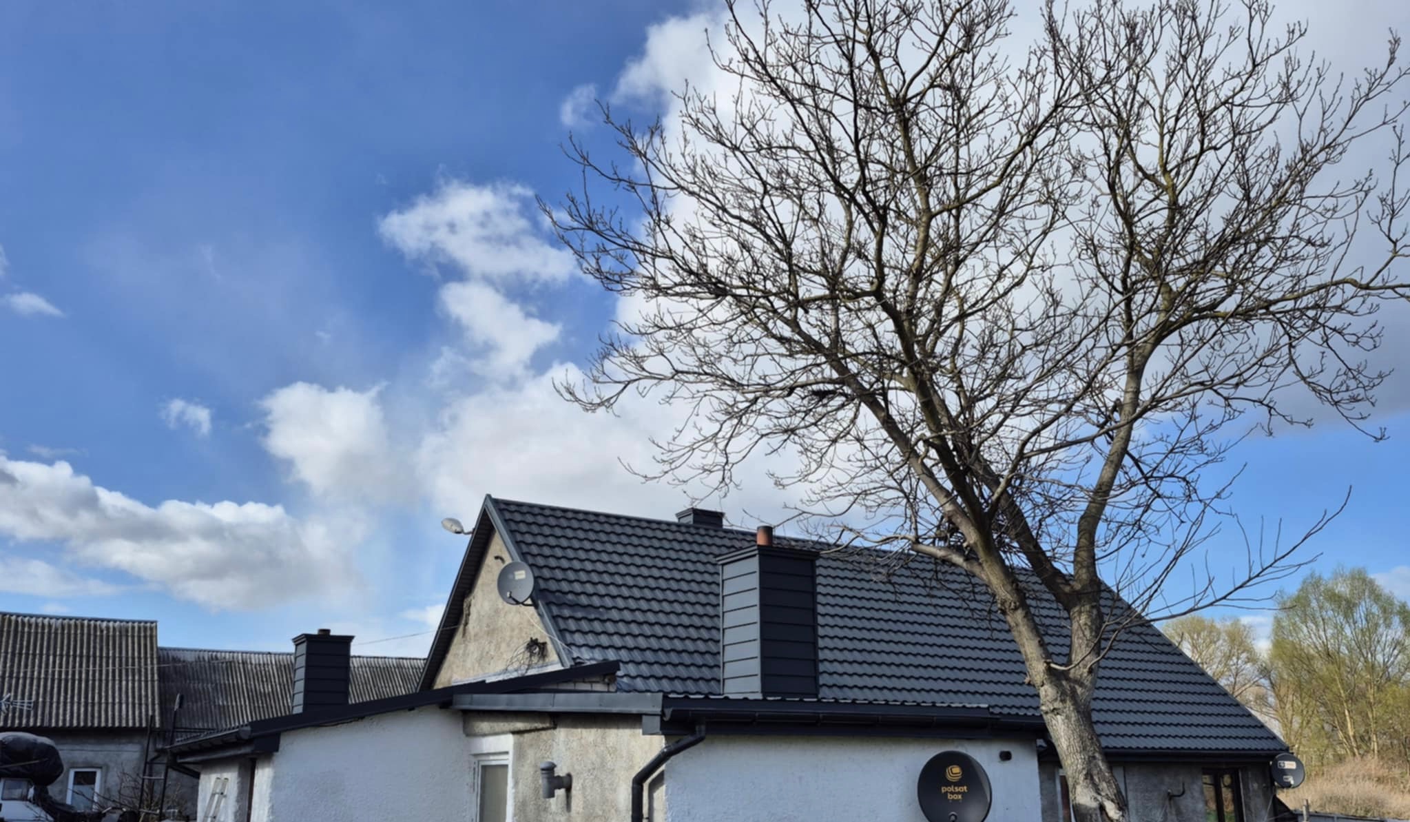 Dom z ciemnoszarą dachówką, widoczny komin obłożony blachą w tym samym kolorze, na ścianie antena satelitarna, na pierwszym planie drzewo bez liści, błękitne niebo z chmurami w tle.
