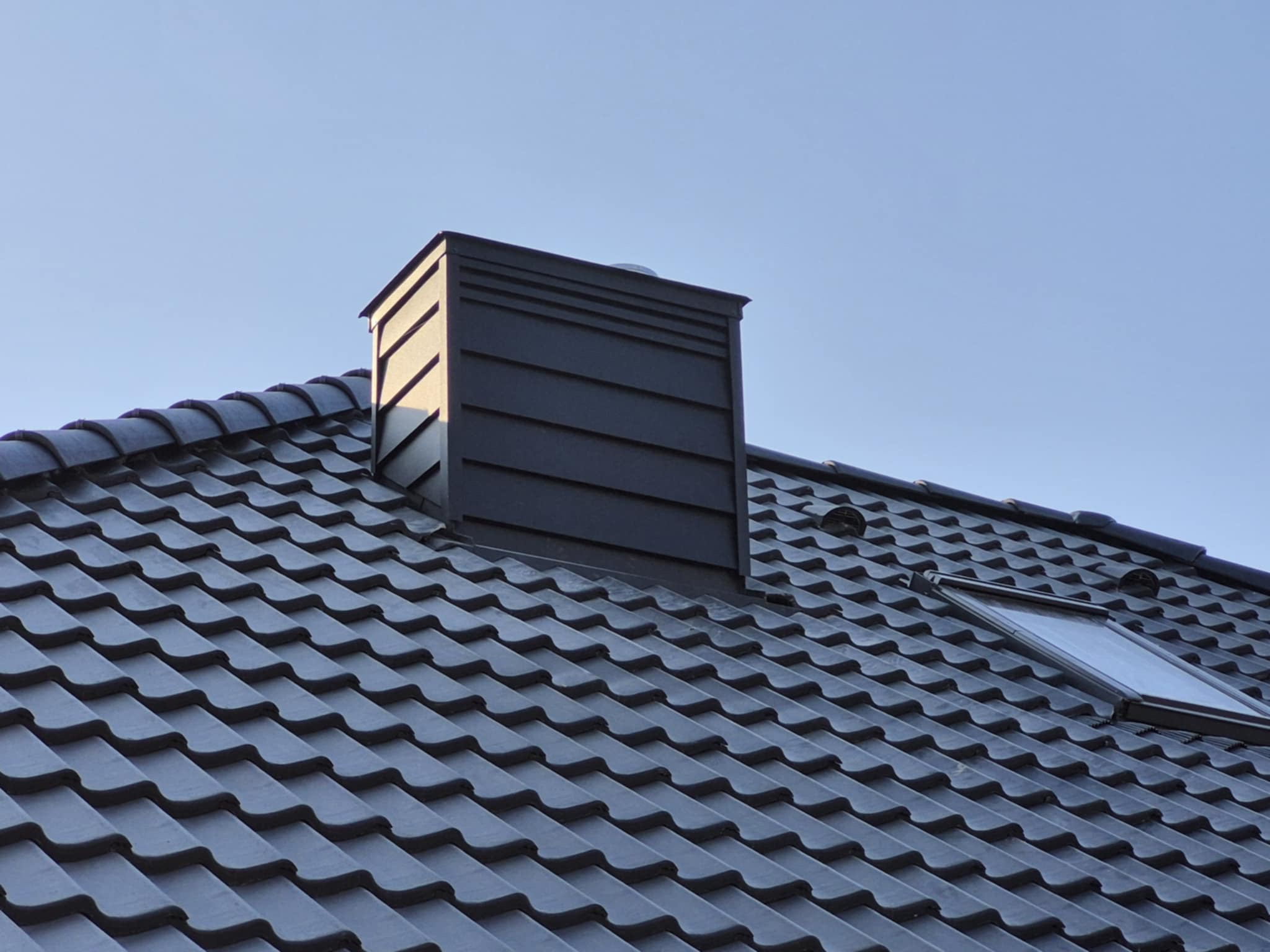 Nowy, ciemnoszary dach z dachówki ceramicznej z oknem dachowym i nowoczesnym kominem w słoneczny dzień.