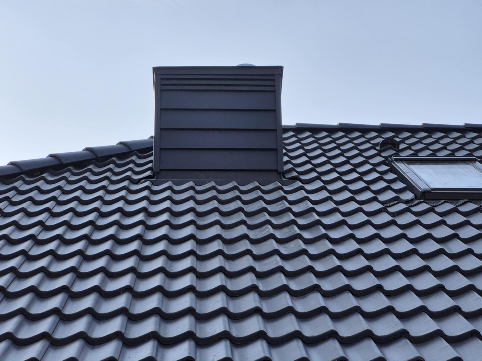 Ciemna dachówka ceramiczna na dachu domu z oknem dachowym i kominem wykończonym blachą w kolorze dachu.