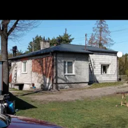 Dwóch dekarzy pracuje na dachu jednopiętrowego domu z ceglanym kominem, widoczne drabiny opierające się o ścianę budynku, częściowo otynkowana elewacja.