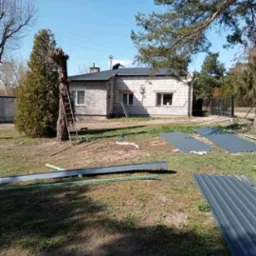 Prace dekarskie w toku: nowy, grafitowy dach z blachy trapezowej na parterowym budynku z białej cegły, dwie drabiny opierają się o dach, na trawniku leżą arkusze blachy i rury PCV.