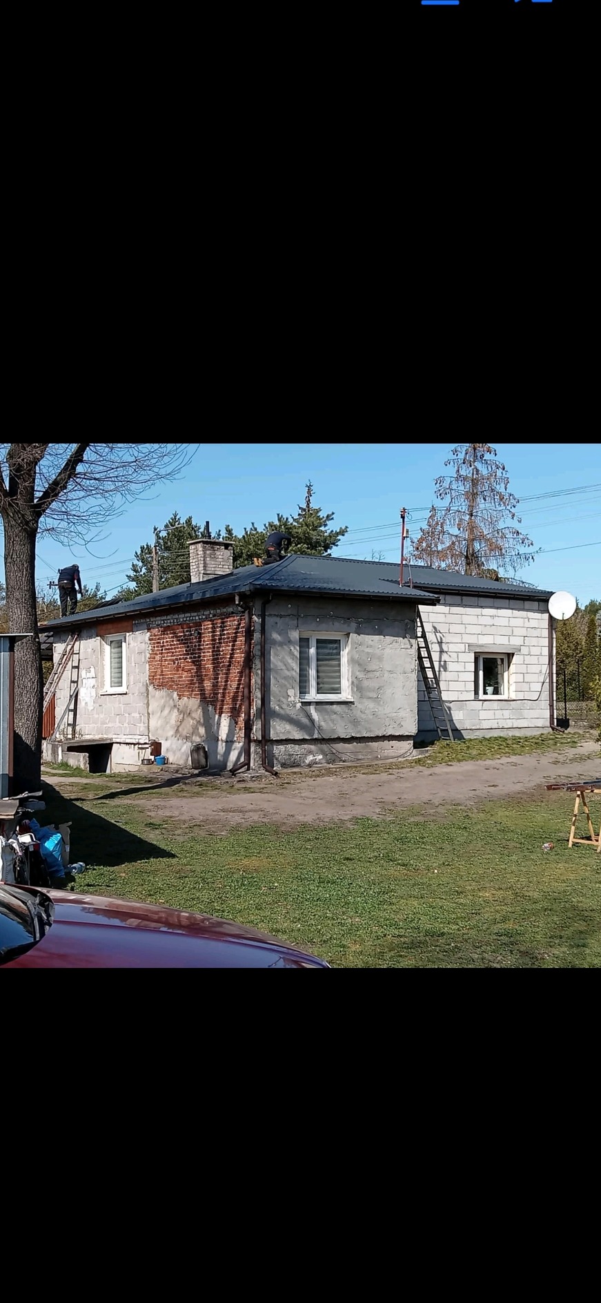 Dwóch dekarzy pracuje na dachu jednopiętrowego domu z ceglanym kominem, widoczne drabiny opierające się o ścianę budynku, częściowo otynkowana elewacja.