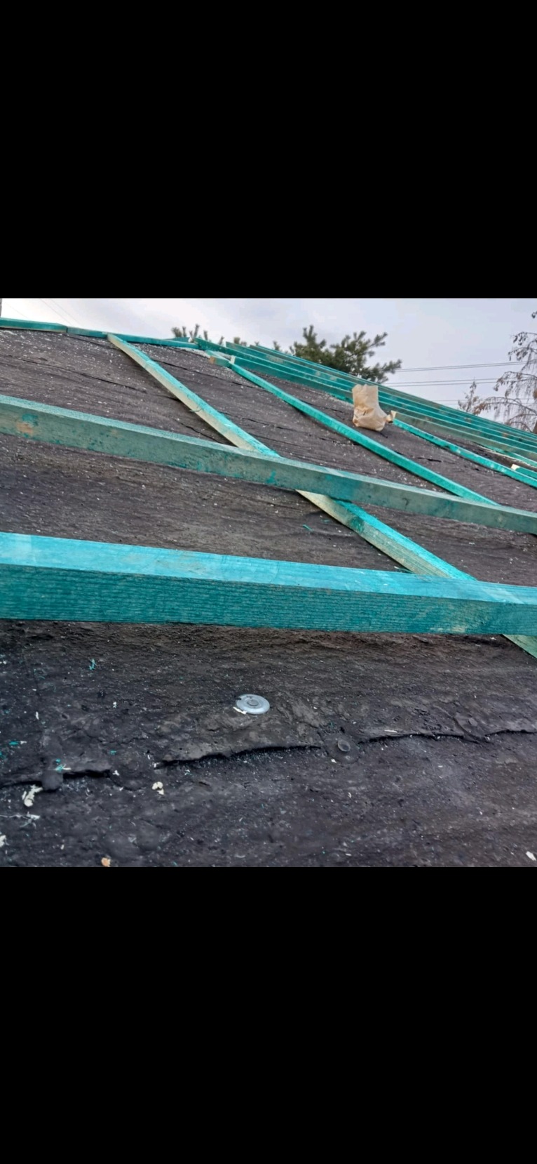 Wstępny etap budowy dachu z widoczną konstrukcją więźby pomalowaną na turkusowo i czarną membraną dachową, zabezpieczoną metalowymi elementami mocującymi.