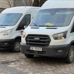 JUK SPÓŁKA Z OGRANICZONĄ ODPOWIEDZIALNOŚCIĄ - Dwa białe busy Ford Transit zaparkowane na brukowanej ulicy, jeden z logo Orlen Paczka na boku, drugi z żółtą kartką za szybą, widoczne numery rejestracyjne.
